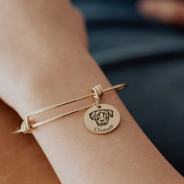 Engraved pet portrait bangle worn on a wrist with a round dog portrait charm and name.