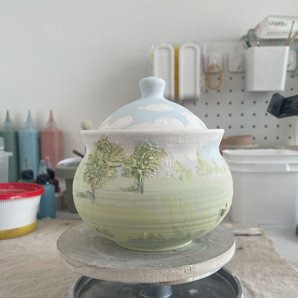Styled rear view of a lidded ceramic pet urn with a soft green landscape glaze and cloud-painted lid.